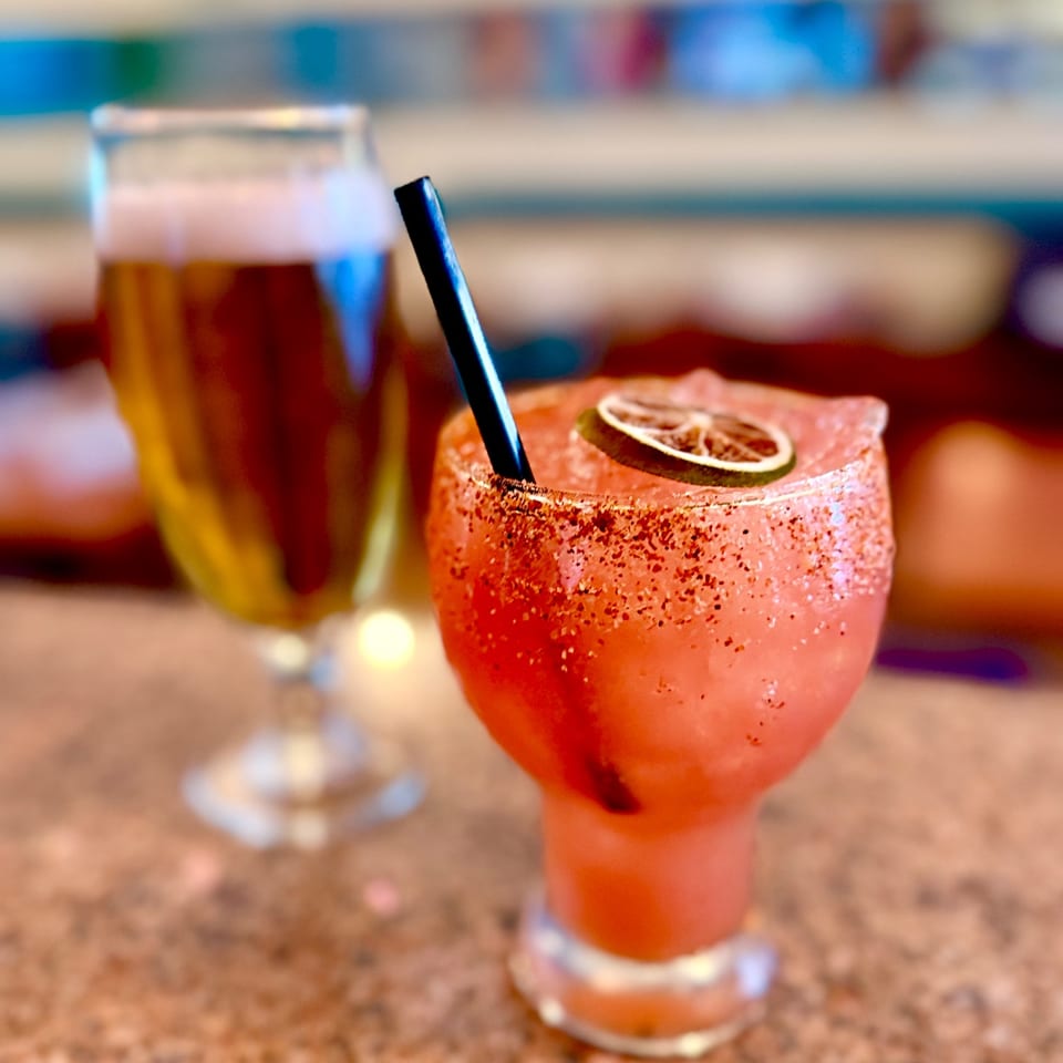 A pink cocktail with a chili rim and lime garnish beside a beer