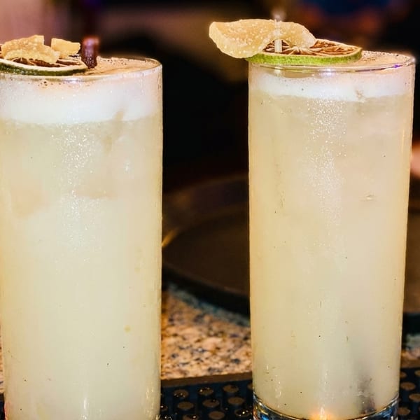 Two tall, frosty cocktails garnished with lime slices and candied ginger on a bar.