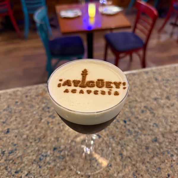 Espresso martini topped with the Ay Guey logo stenciled in cinnamon, on the bar with the colorful dining room behind.